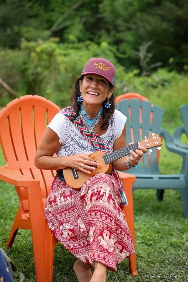 A woman in a Barefoot Circus cap playing a ukulele in a lawn chair
